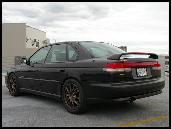 Black 1998 Subaru Legacy sedan with a rear spoiler and bronze wheels, viewed from the rear three-quarter angle