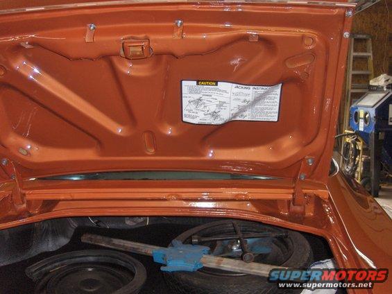 Car trunk open showing spare tire, jack, and caution sticker on the underside of the trunk lid