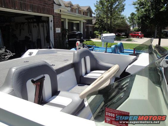 Boat interior with two-tone seats, steering wheel, and dashboard gauges. Mercury outboard motor visible in background. Red car in driveway