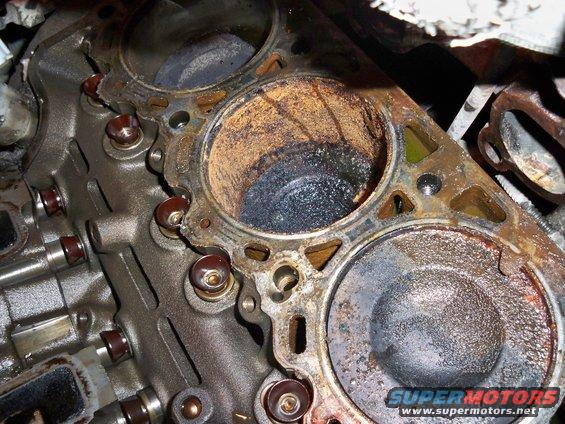 Close-up of a damaged engine cylinder with carbon buildup and rust. The cylinder head is removed, showing the piston top and surrounding metal
