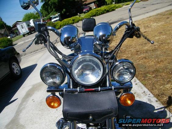 Motorcycle front end with dual headlights and chrome accents. Black leather pouch on handlebars