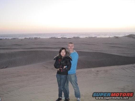 Couple posing on sand dunes with the ocean and distant RVs in the background