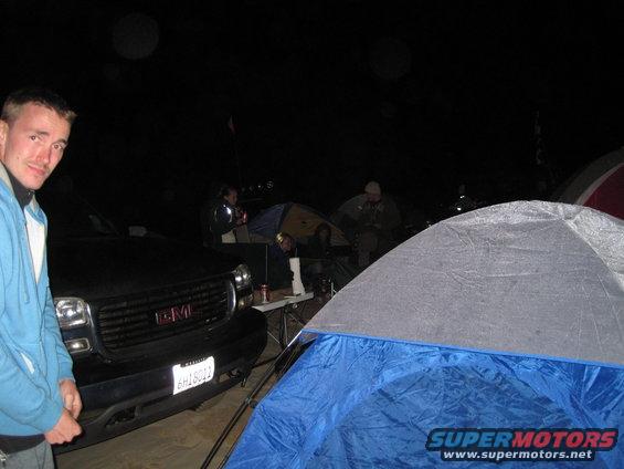 Guy in blue jacket next to black GMC Sierra pickup truck at campsite with tents and people