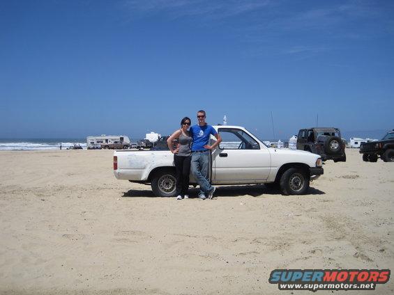 couple by white pickup truck on sandy beach, other vehicles and RVs in background