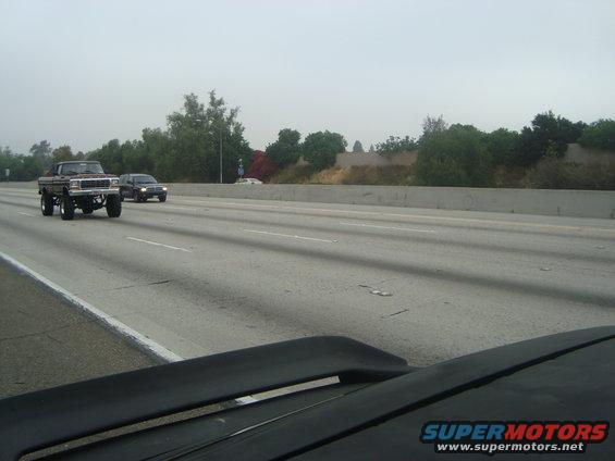 Brown lifted pickup truck driving on highway, front 3/4 view. Other vehicles and trees visible