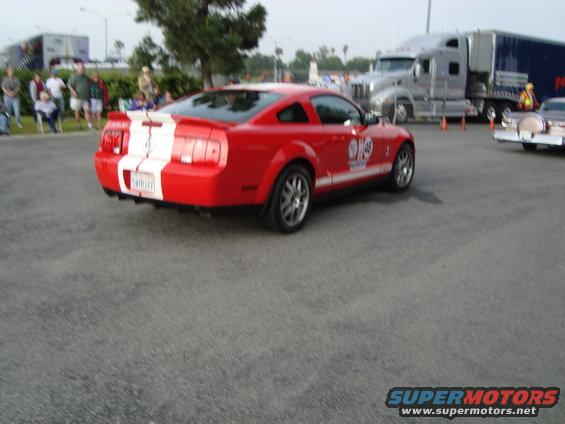 Red Ford Mustang with white racing stripes on rear and side, seen from rear quarter