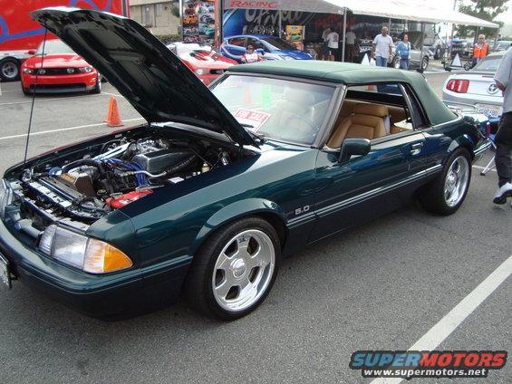 Green convertible Mustang with hood open, showing engine. Other cars and tents in background