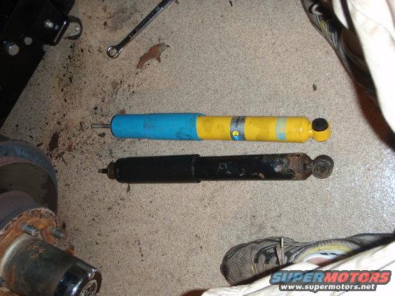 Two shock absorbers, one yellow and blue, the other black and rusty, lie on a concrete floor next to a car part