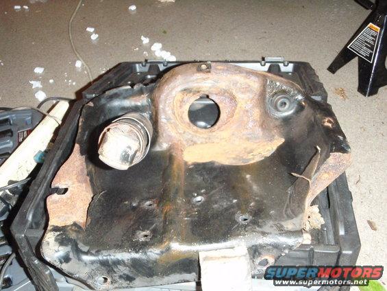 heavily rusted metal car part with a circular opening and a pipe sticking out, sitting on a black plastic container
