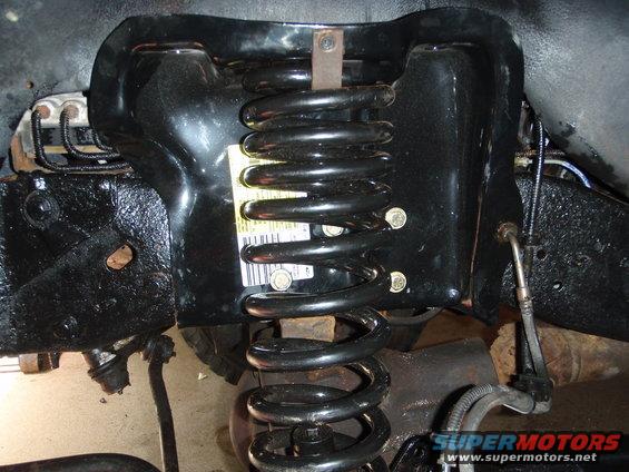 Close-up of a black coil spring attached to a black metal mount, part of a vehicle's front suspension system