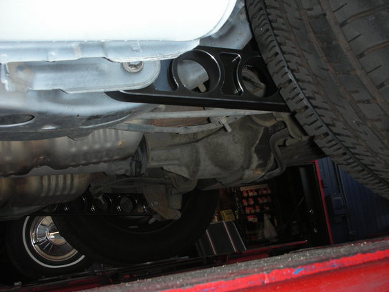 Underside view of a car's rear suspension with a black aftermarket control arm installed. Tire and differential visible