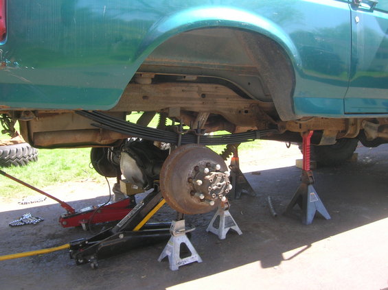 Green truck's rear axle removed, supported by jack stands. Leaf springs and brake drum visible