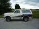 1988 Ford Bronco - White and blue Ford Bronco parked on asphalt with a rural landscape in the background.