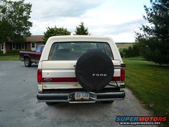 Rear view of a white Ford Bronco with a spare tire cover and a red stripe. A red pickup truck is parked nearby