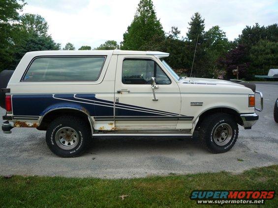 White and blue Ford Bronco with rust spots and a spare tire on the back, viewed from the side