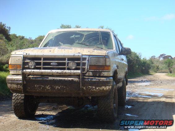 1992 Ford Bronco covered in mud, parked on a dirt road with trees and sky in background