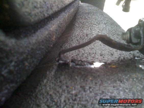 Close-up of a dark, textured vehicle part with a bent wire and a bolt. A white substance is visible near a crack