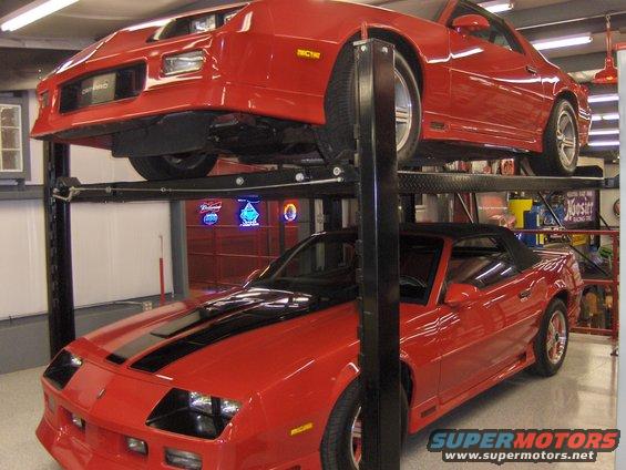 Two red Camaros stacked on a car lift. The bottom one is a convertible