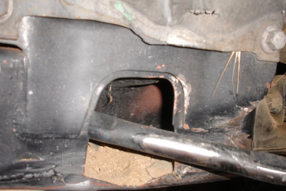 Black metal vehicle subframe with a hole revealing a rusty pipe, viewed from below