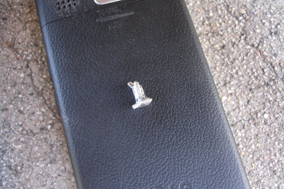 Small, silver, T-shaped metal object with a loop on a black, textured surface, possibly a phone back