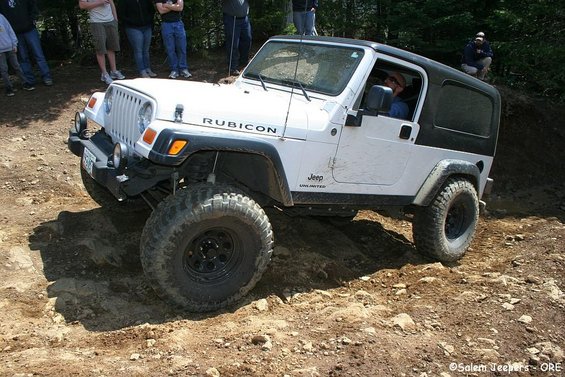 White Jeep Wrangler Rubicon on a dirt trail, front 3/4 view. Muddy tires and body