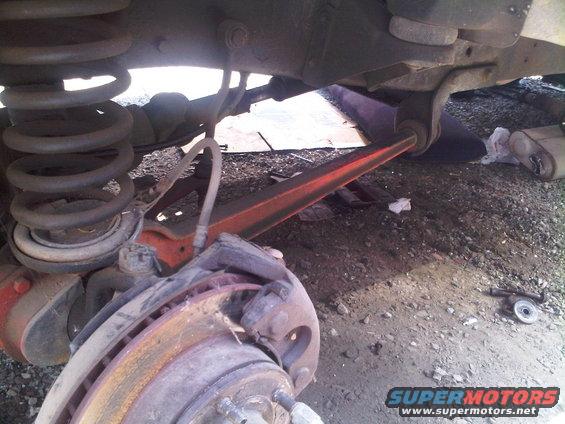 Close-up of a vehicle's rear suspension, showing the coil spring, brake rotor, and control arm. The vehicle appears to be on uneven ground, possibly a junkyard