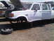 1986 Ford Bronco - Junkyard scene shows a white Ford F-350 truck missing its front wheels and with its hood open.