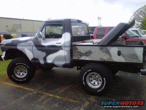 Camouflaged pickup truck with a flatbed and a red truck behind it