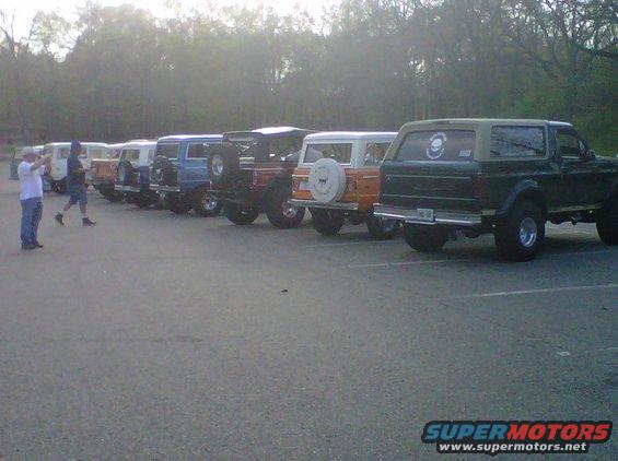 Line of Ford Broncos parked, rear view of green 1995 Ford Bronco prominent. Two people stand near the front