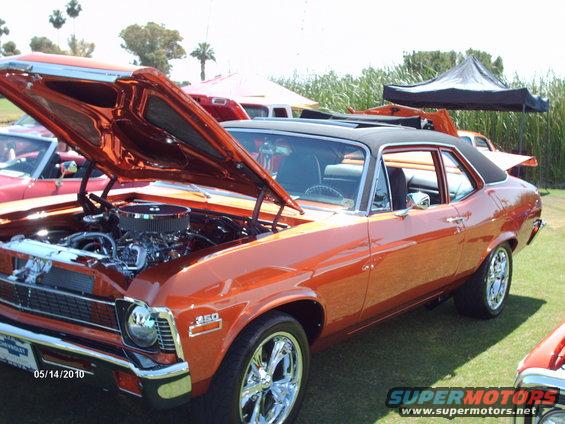 Orange 1970 Chevrolet Nova with hood open, showing engine. Another red car is partially visible to the left