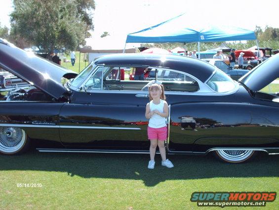 Black coupe with hood open, girl standing beside it