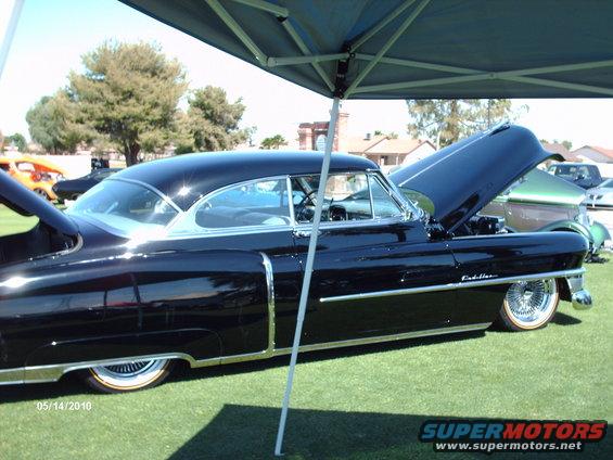 Black 1950s Cadillac with hood open, parked on grass under a canopy. Another car is visible behind it