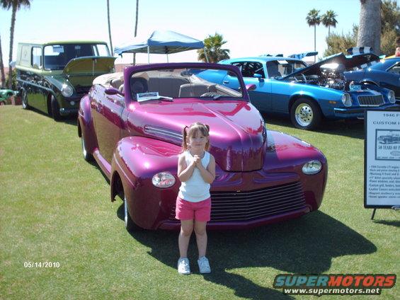 Purple convertible with a young girl standing in front. Other cars and a sign are visible