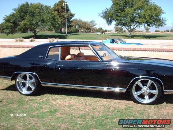 Black coupe with tan interior and chrome wheels parked on grass, with another blue car in the background
