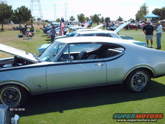 Silver coupe with black stripe, hood up, parked on grass at car show. Other cars and people in background