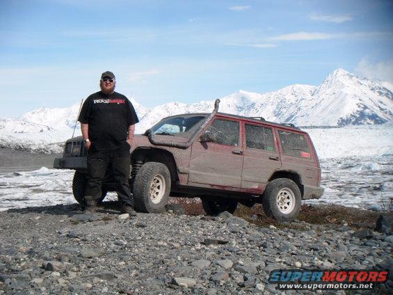 Man standing next to a red 1998 Jeep Cherokee with a snorkel in a rocky, icy landscape