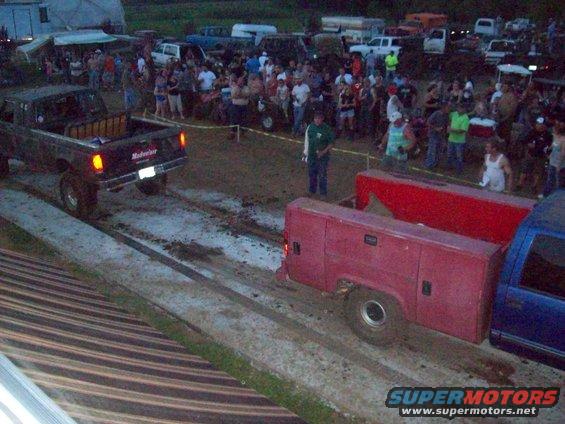 Black pickup truck pulling a red trailer with toolboxes, crowd watching in background