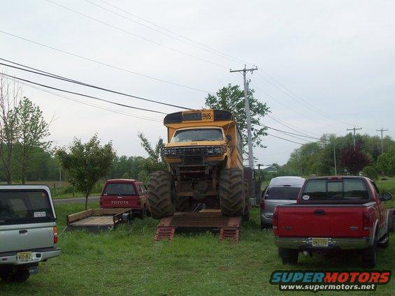 Yellow school bus body on monster truck chassis with huge tires. Other trucks and a trailer are parked nearby