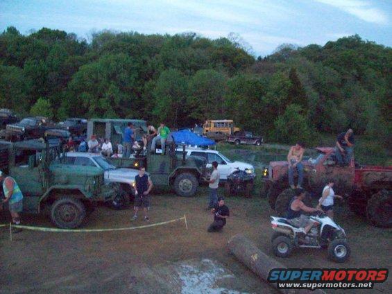 Muddy scene with various trucks, a white SUV, an ATV, and people gathered