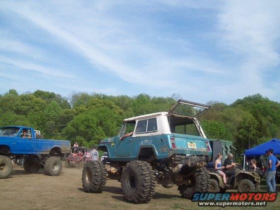 Blue and white Jeep CJ-5 with oversized tires, rear view. Blue pickup truck and ATVs also visible