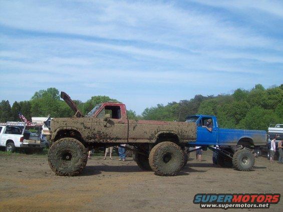 muddy lifted pickup trucks side by side, one red and brown, one blue. Hood is up on the red truck