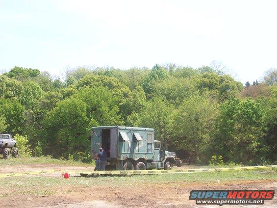 Military truck with a box body parked in a grassy area with trees behind it. Another vehicle with large tires is visible to the left