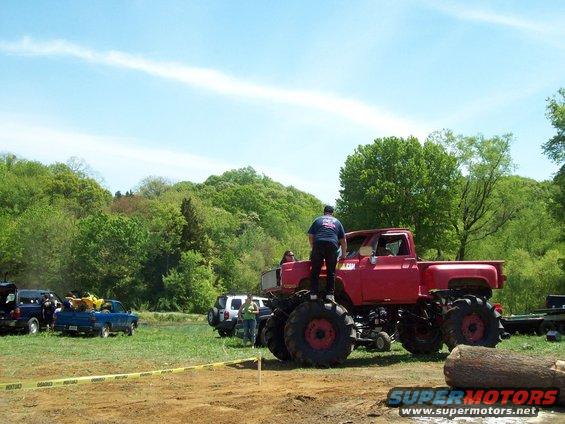 Red monster truck with oversized tires, people standing on the hood and roof, in a muddy outdoor area with other vehicles