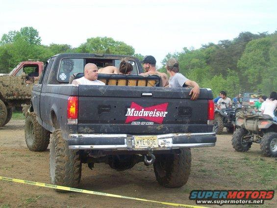 Rear view of a black pickup truck with a 'Mudweiser' sticker on the tailgate, carrying passengers. Another muddy truck is visible to the left, and ATVs are in the background