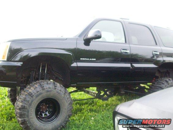 Lifted black SUV with large tires, side view. Another vehicle is partially visible in the foreground