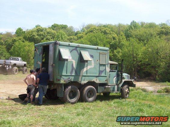 Camouflaged military truck with people loading, monster truck in background