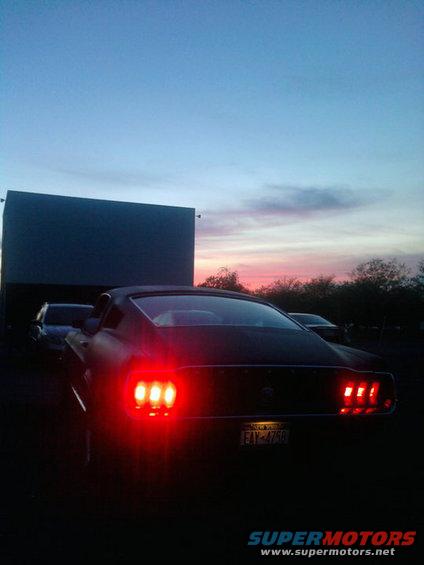 Rear view of a dark-colored classic Mustang with its brake lights on, parked in front of a drive-in movie screen at dusk. Another car is visible behind it