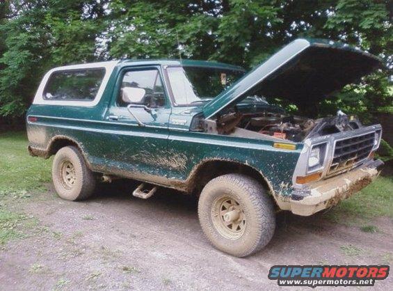 Green 1970s Ford Bronco with hood open, covered in mud. Front grille and bumper are also muddy