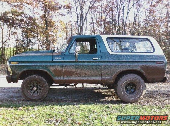 Green and white Ford Bronco parked on dirt road with trees in background