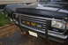 1988 Ford Bronco - The front grille and headlights of a dark blue Ford Bronco are visible, with a chrome bull bar and yellow fog lights.